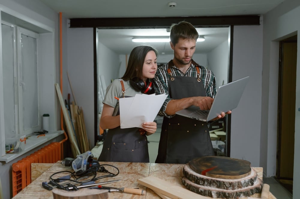 Zwei Personen in Arbeitskleidung stehend in einer Werkstatt mit Werkzeugen und Holz. Sie halten Papier und einen Laptop in den Händen als Symbol der Digitalisierung im Handwerk.